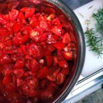 Im Vordergrund eine große Metallschüssel mit klein geschnittenen Tomaten. Daneben liegen ein paar Zweige Rosmarin, die gerade gezupft werden.
