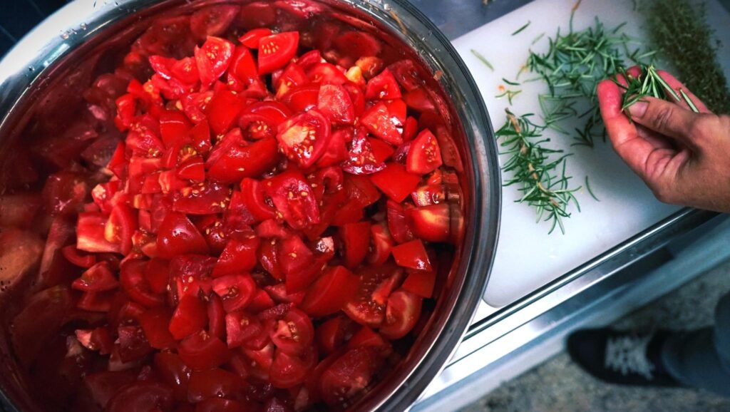 Im Vordergrund eine große Metallschüssel mit klein geschnittenen Tomaten. Daneben liegen ein paar Zweige Rosmarin, die gerade gezupft werden.