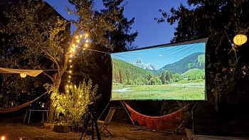 Leinwand hängt in einem Garten zwischen Obstbäumen. Auf der Leinwand ist eine Berglandschaft.